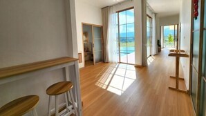 Amplia habitación con suelo de madera, un gran ventanal corredero que da a un balcón y una pequeña zona de estar con taburetes.