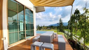Un patio exterior moderno con piso de madera, asientos grises y un amplio ventanal que da a una piscina.