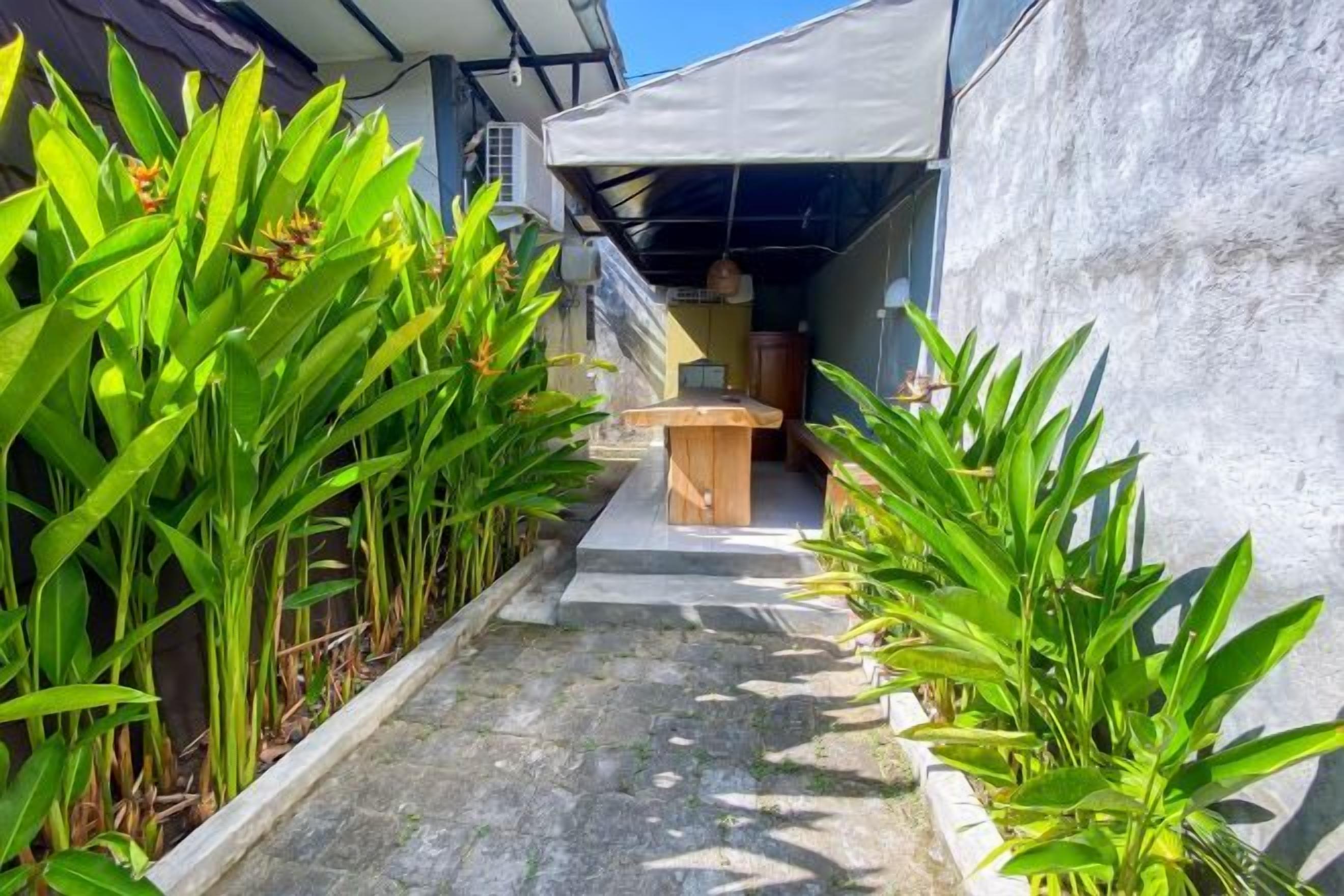 A narrow pathway flanked by green plants leading to a covered outdoor area with a table and chairs.