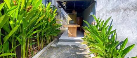 A narrow pathway flanked by green plants leading to a covered outdoor area with a table and chairs.