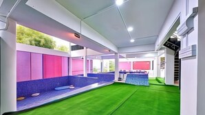 A modern indoor pool area with a green artificial turf floor, a seating area with blue and pink cushions, and a blue pool table. - Penang Dream Banglo (Tanjung Bungah)