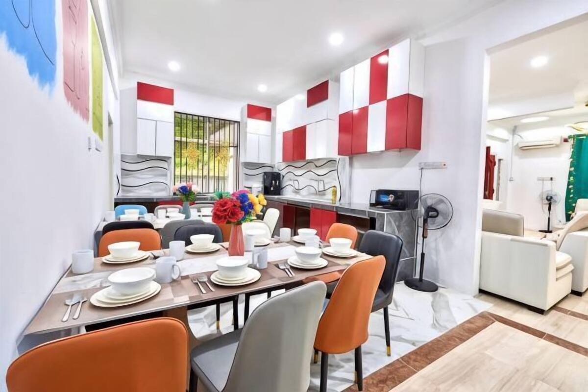 A modern dining area with a table set for four, a vase with flowers, and a kitchen in the background.
