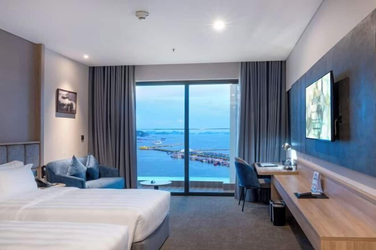 A hotel room with a bed, a desk, a chair, a TV, and a large window with a view of the sea and buildings.