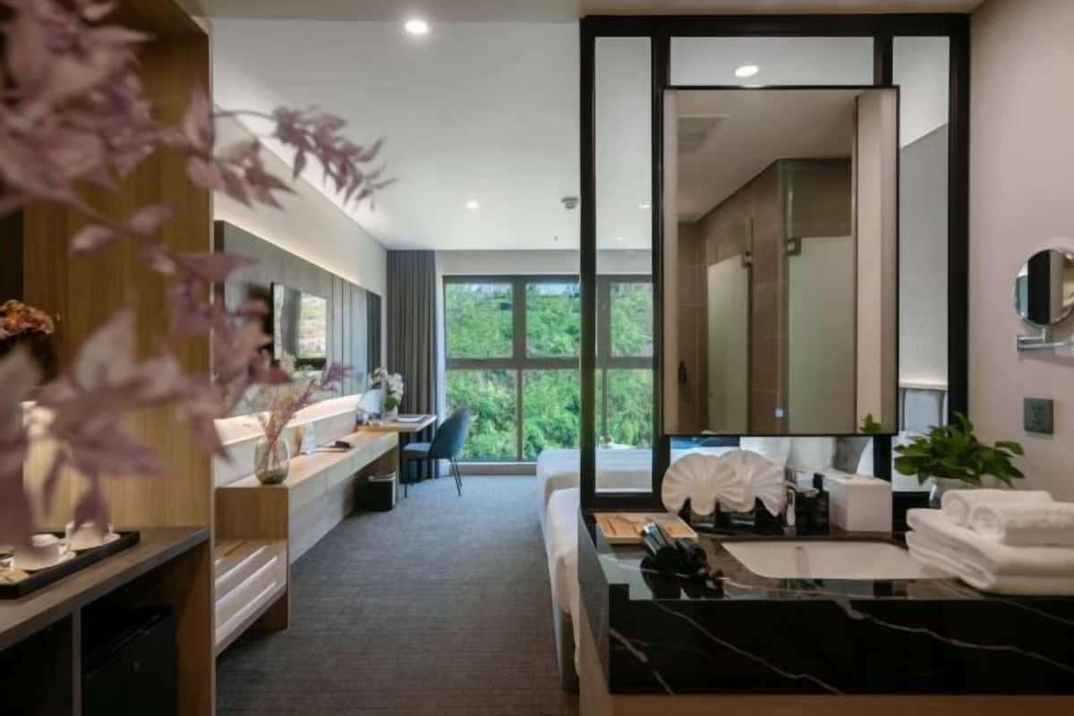 A modern hotel room with a large bed, a desk, a bathroom with a large mirror, and a view of greenery outside.