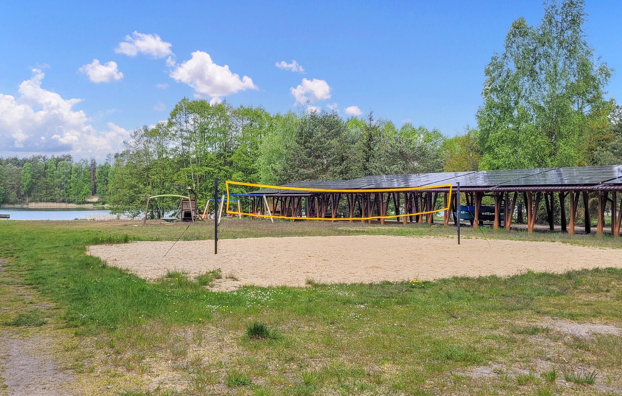 A beach volleyball court with a net, surrounded by sand and grass, near a lake and trees.