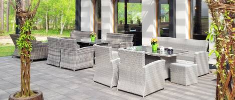 En udendørs terrasse med fletmøbler, et glasbord og udsigt til en skov.