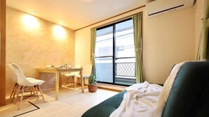 A small, well-lit room with a bed, a desk, and a chair. There is a window with curtains, a potted plant, and an air conditioning unit on the wall.