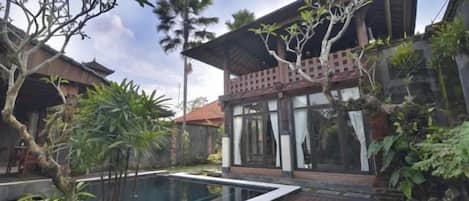 A pool with a wooden deck, surrounded by tropical plants and a building with large windows and a balcony.