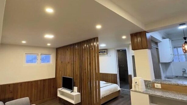A modern hotel room with a wooden panel wall, a bed, a TV, a small seating area, and a kitchenette. - Thamel Holiday Apartments (Kathmandu)