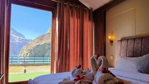 A hotel room with a bed, a window with curtains, a view of mountains, and a towel rack with towels.