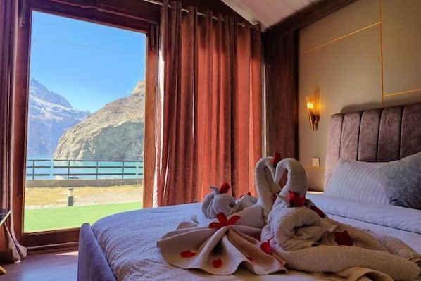 Un cuarto de hotel con cama, ventana con cortinas y vistas a montañas, además de un perchero con toallas.