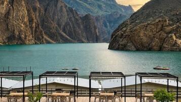 A lakeside terrace with outdoor seating and a view of a mountainous landscape.