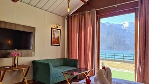 A bedroom with a bed, a green sofa, a small table, a TV, and a view of mountains and a body of water.