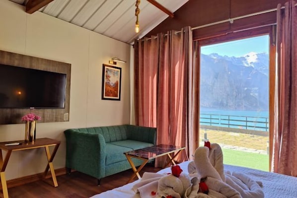 A bedroom with a bed, a green sofa, a small table, a TV, and a view of mountains and a body of water. - Horizon Resort Hunza (Gilgit)