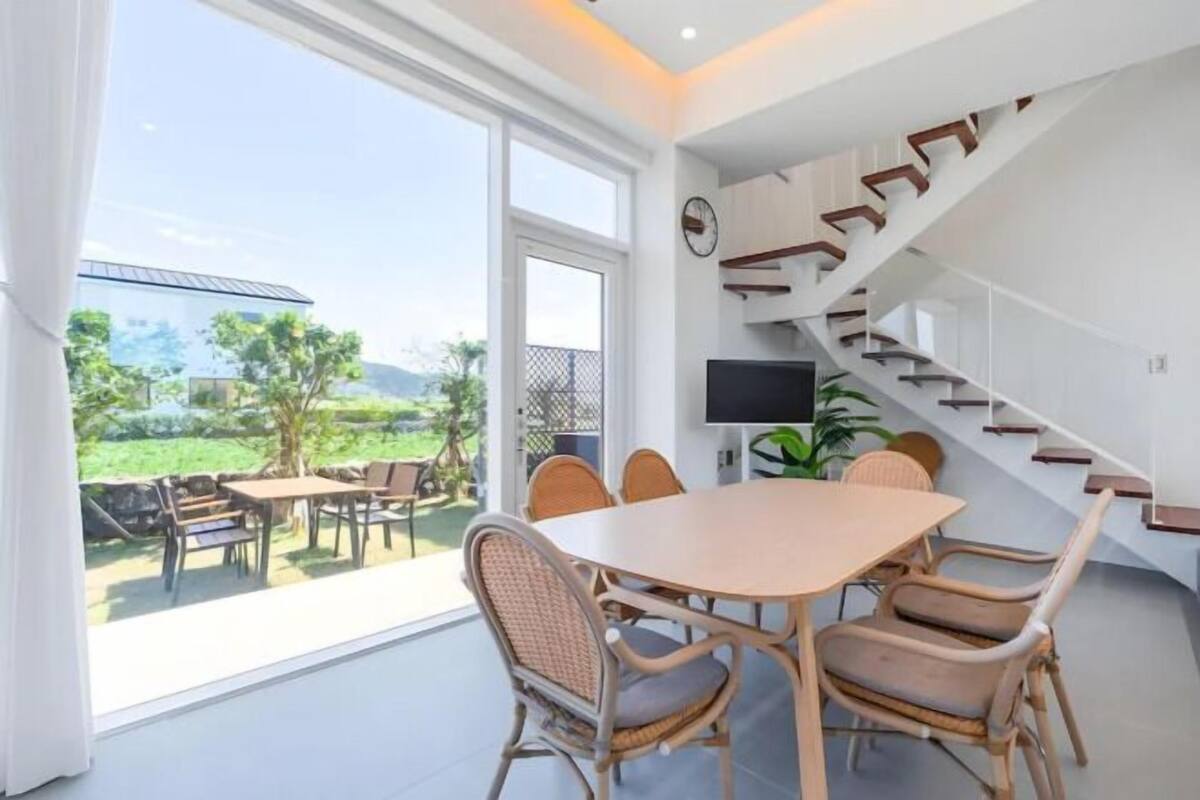 A modern interior with a dining area, a staircase, and a large window offering a view of the outdoors.