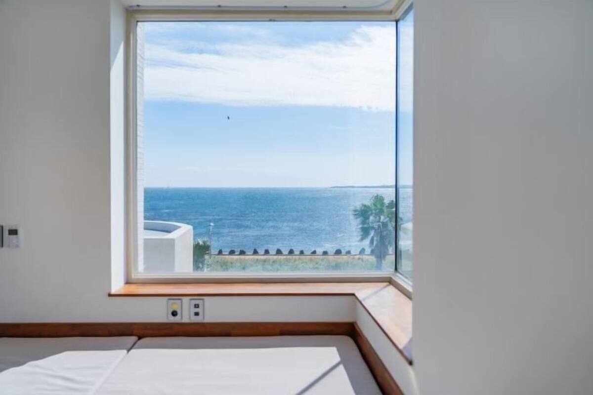 A room with a large window overlooking a body of water, a bed, and a small wooden bench.