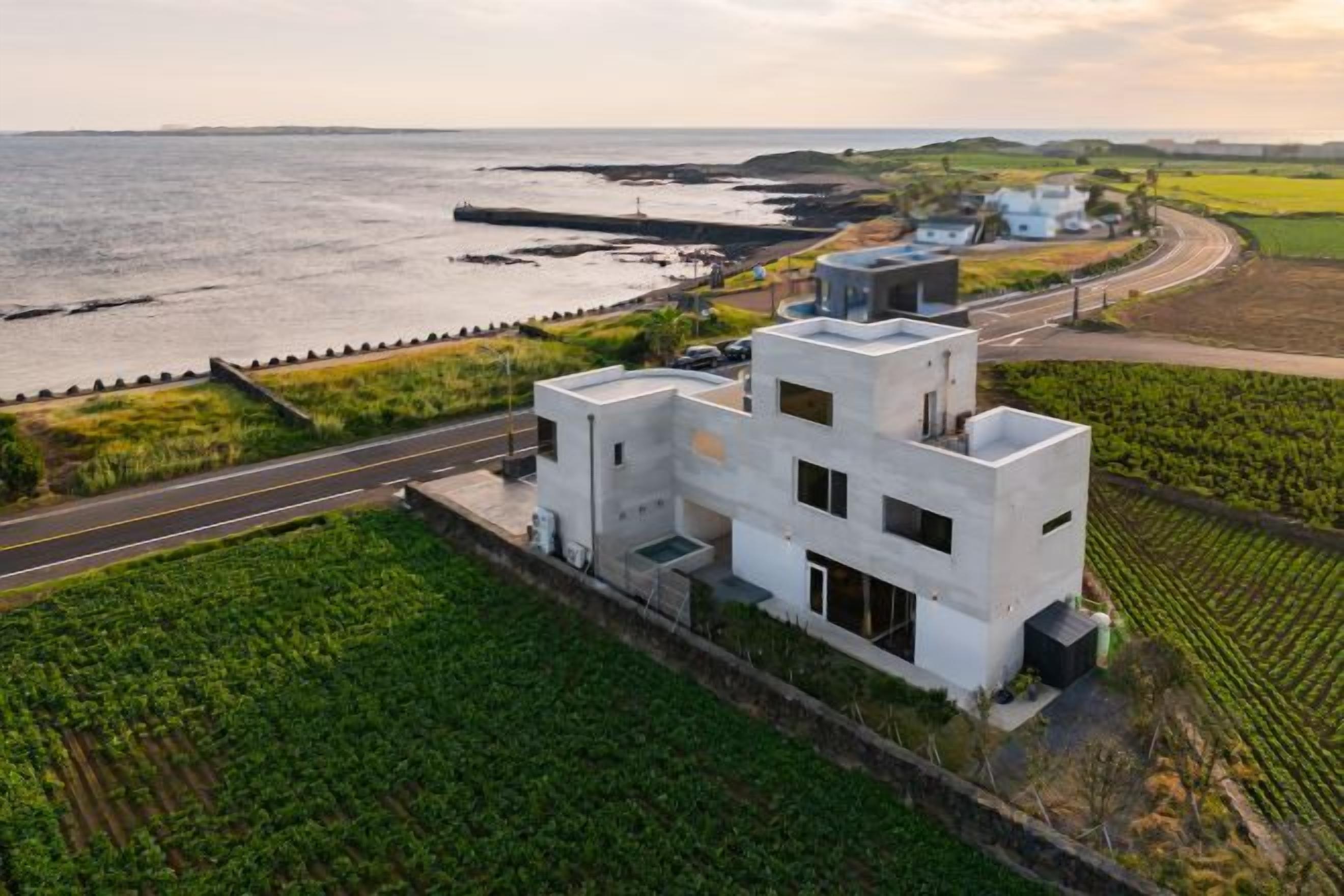 A modern white building with a flat roof, located near a body of water and a road.