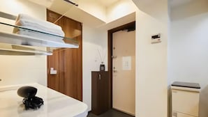Habitación de hotel con encimera blanca, un objeto negro encima, estante de vidrio con toalla, puerta de madera y un mueble pequeño.