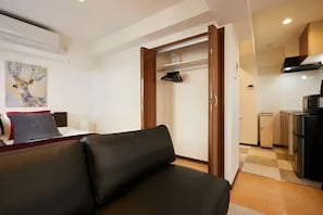 Moderni hotellihuone, jossa on musta nahkasohva, puinen vaatekaappi ja taustalla pieni keittiö.