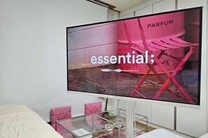 Un televisor mostrando un anuncio de una marca de perfume, con una silla rosa y una pared de ladrillo al fondo.