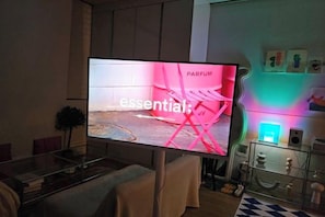 Un televisor de pantalla plana montado sobre un soporte en una sala de estar con una mesa de centro de vidrio, un sofá y una planta decorativa.