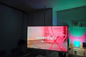 Un televisor mostra un anunci de 'PARFUM essential' en una habitació amb un disseny modern, inclosa una taula de vidre i una estant amb objectes.