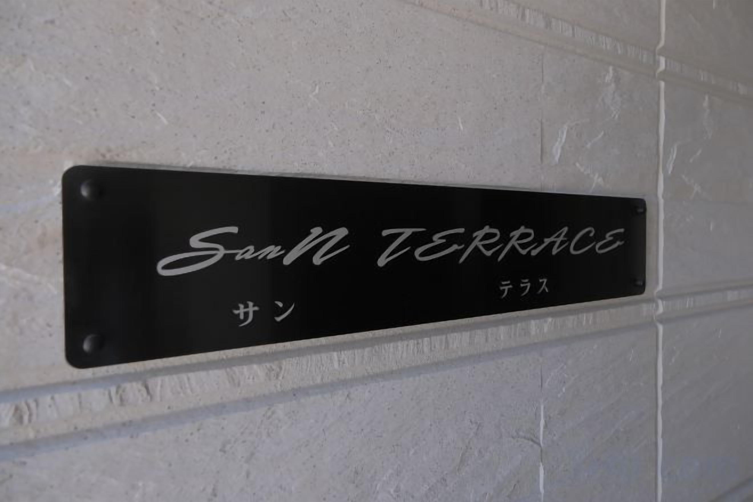 A black metal sign with white lettering on a white wall, reading 'San TERRACE' with additional text in another language below.