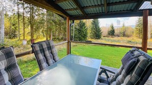 A covered outdoor seating area with a glass table and plaid cushions, overlooking a grassy landscape with trees. - 2 bedroom gorgeous home in Rottneros (Rottneros)