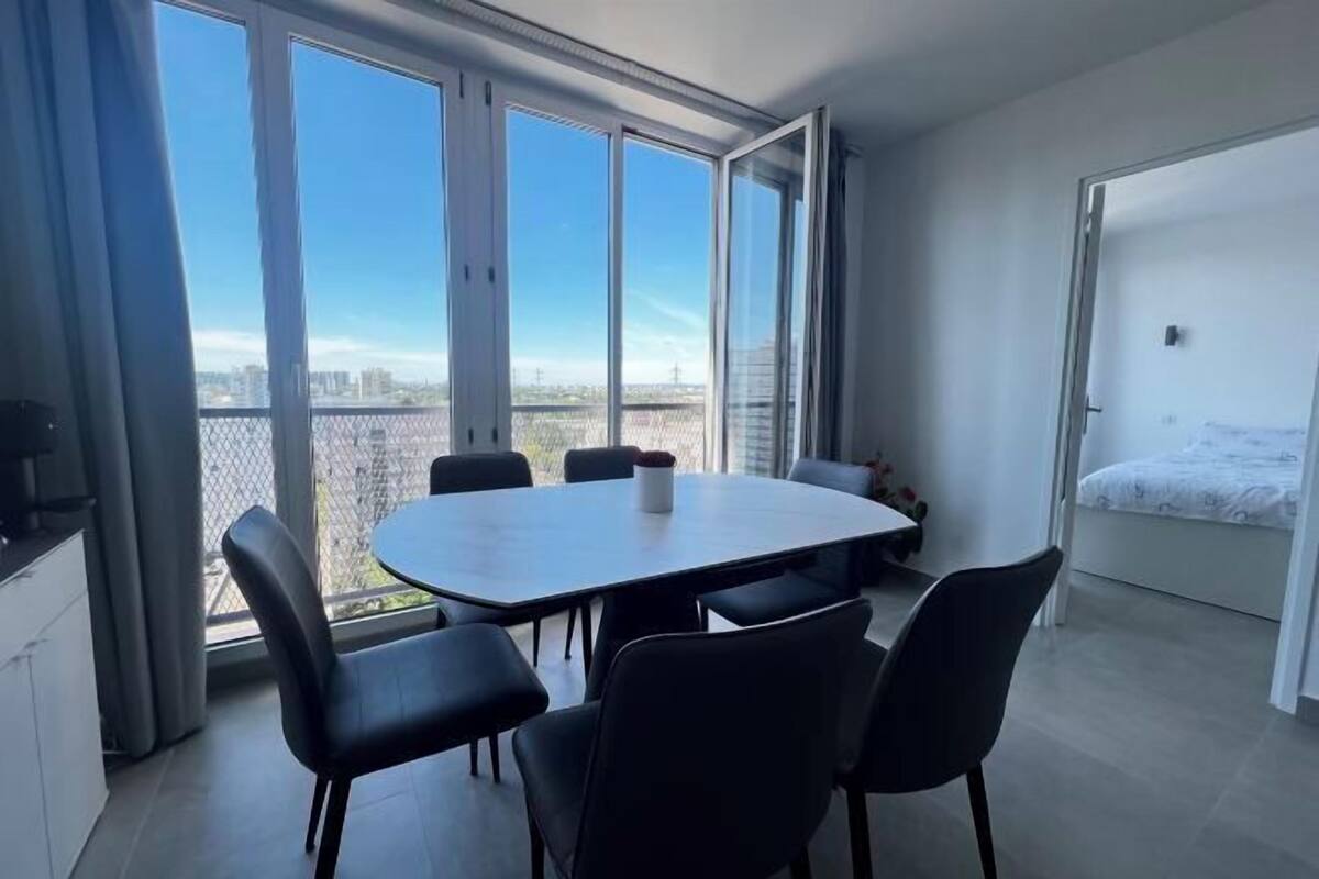 Una habitación moderna con un amplio ventanal, una mesa de comedor con sillas, una cama y una pequeña cocina.