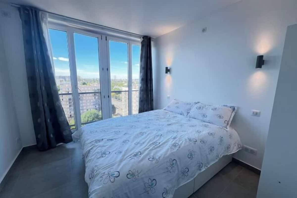 A bedroom with a bed, a window with curtains, a wall-mounted light, and a view of the city.