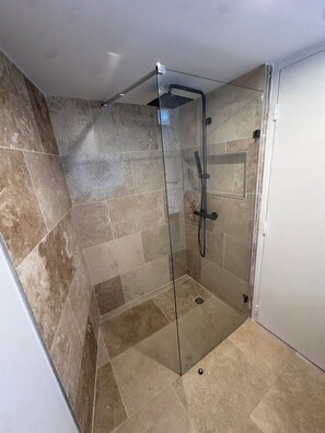 Una ducha con mampara de vidrio, cabezal de ducha de mano, paredes con azulejos y desagüe en el suelo.