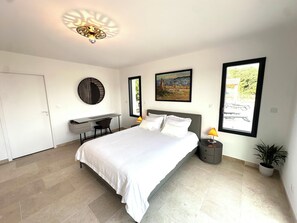 A bedroom with a bed, desk, chair, nightstand, lamp, and a potted plant.