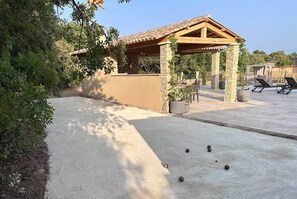 A covered outdoor seating area with stone walls and a tiled roof, surrounded by greenery and potted plants.