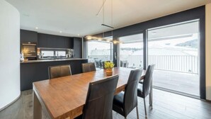 A modern dining area with a wooden table and chairs, a kitchen in the background, and large windows offering a view of a cityscape.