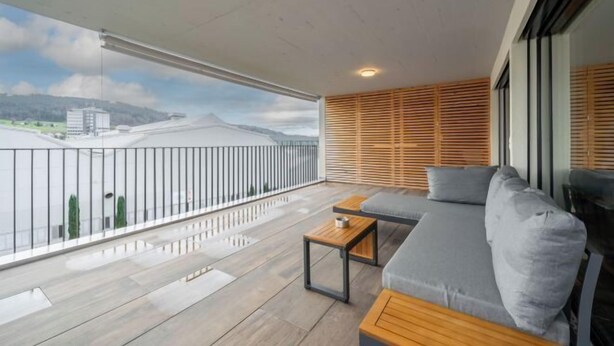 A balcony with a grey sofa, wooden table, and a view of a cityscape.