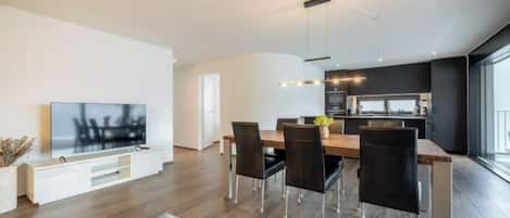 Un salon moderne doté d’un téléviseur à écran plat, d’un coin repas avec des chaises et d’une cuisine en arrière-plan.