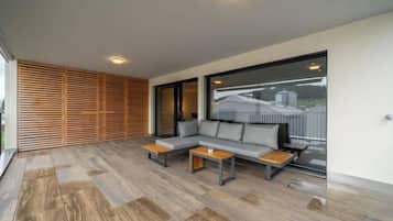 A modern outdoor seating area with a sofa and two tables, a wooden slatted wall, and a large window with a view.