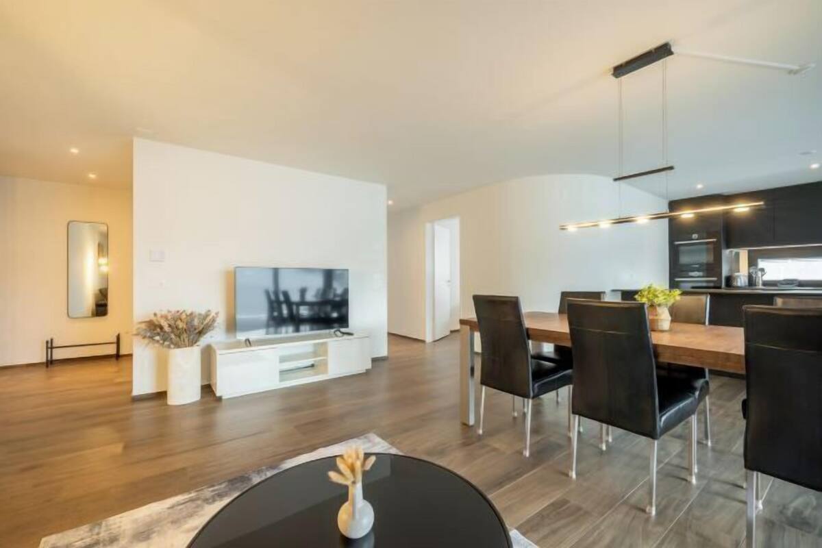 A modern living room with a flat-screen TV, dining area with chairs, and a kitchen with a countertop.