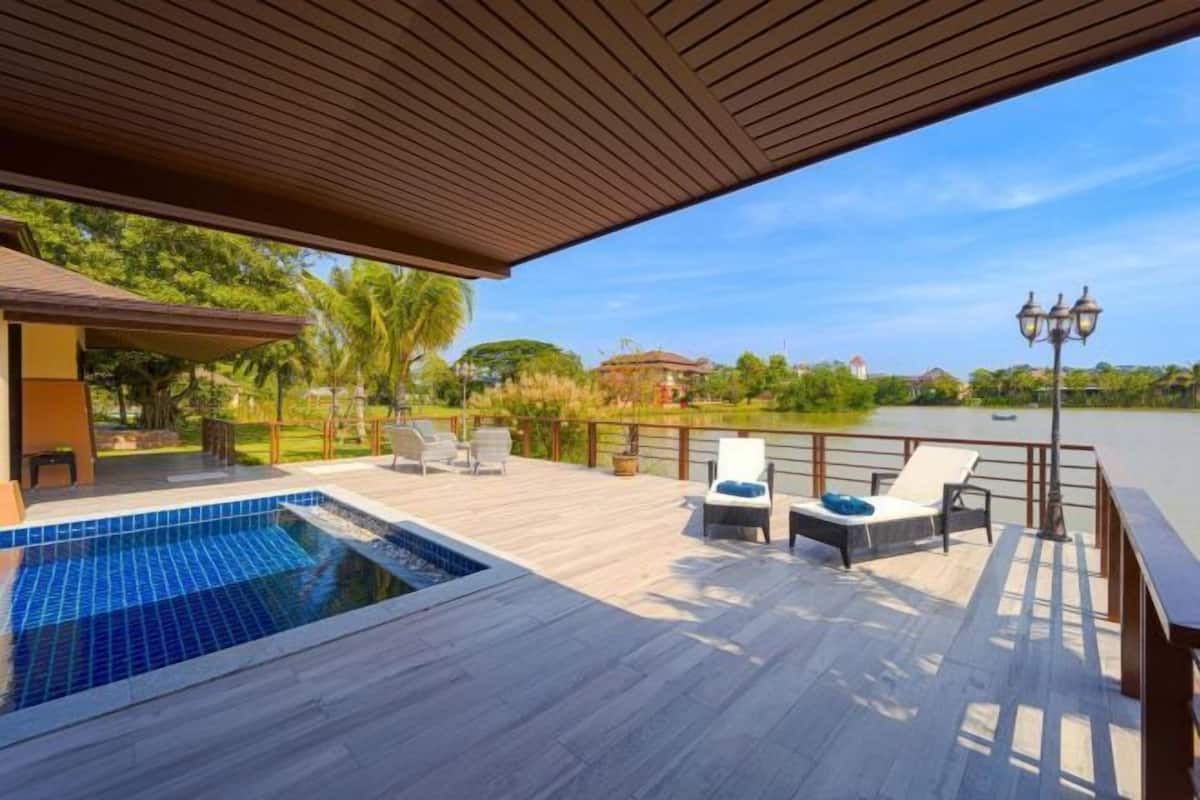 A pool area with a wooden deck, lounge chairs, and a view of a lake.