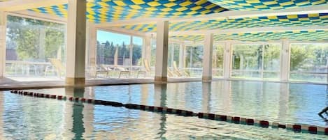 Une piscine intérieure à l’eau claire, avec un plafond à carreaux et une vue sur les arbres à l’extérieur.
