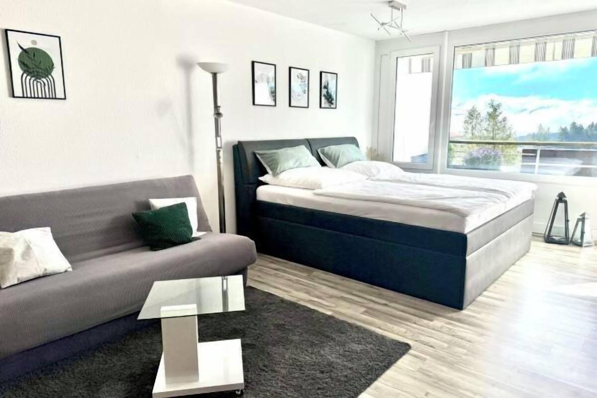 A modern bedroom with a grey sofa, a bed with dark headboard, a small table, and a large window with a view.