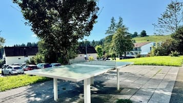 Uma mesa de pingue-pongue ao ar livre em um parque, com área pavimentada, cercada por árvores e um bairro residencial ao fundo.