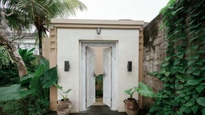 A white door with a decorative arch, flanked by two potted plants, leading to a building with a stone wall covered in ivy. - Sapta Villa Ubud (Bali)