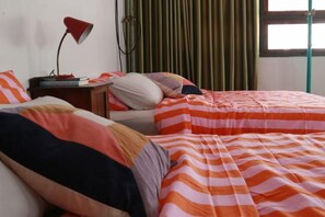 A bedroom with two beds, striped bedding, a nightstand with a lamp, and a window with curtains. - The Urbanists BKK (Bangkok)