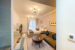A modern hotel room with a bed, a sofa, a round coffee table, a desk with a lamp, and a ceiling light fixture. - Cosmos Living Luxurious Studio Near Dubai Marina (Dubai)