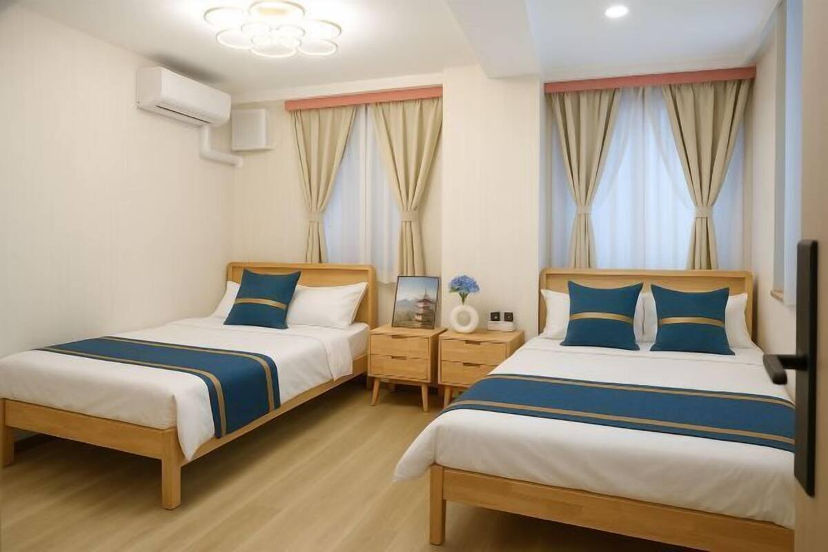 A hotel room with two beds, wooden headboards, beige curtains, a nightstand with a vase, and an air conditioning unit.