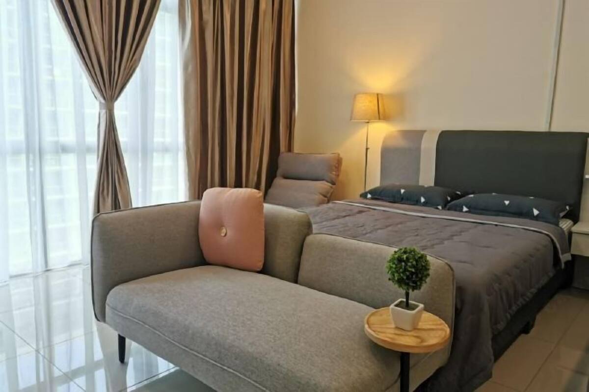 A modern hotel room with a sofa, a bed with a dark headboard, a bedside lamp, and a small potted plant.