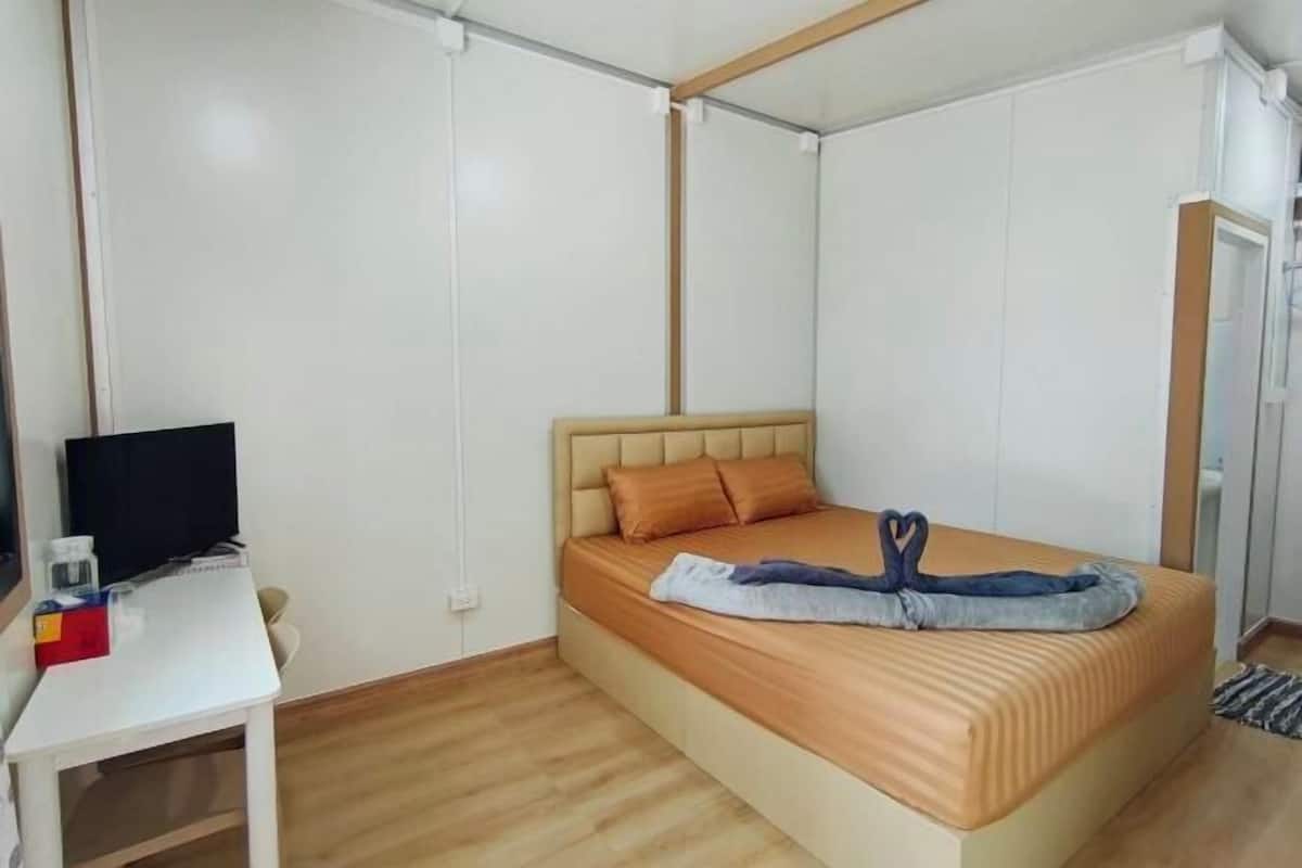 A single bed with a headboard, a bedside table with a television, and a small table with a box and a bottle.