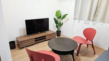 A small living area with a flat-screen TV on a wooden stand, a round table, two red chairs, and a potted plant.