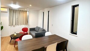 A modern room with a wooden dining table, red and white chairs, a grey sofa, and a window with a black frame. - Yahiro 6chome (Tokyo)